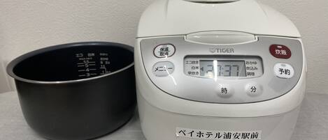 A white rice cooker with a digital display and buttons in Japanese, next to a black measuring cup.