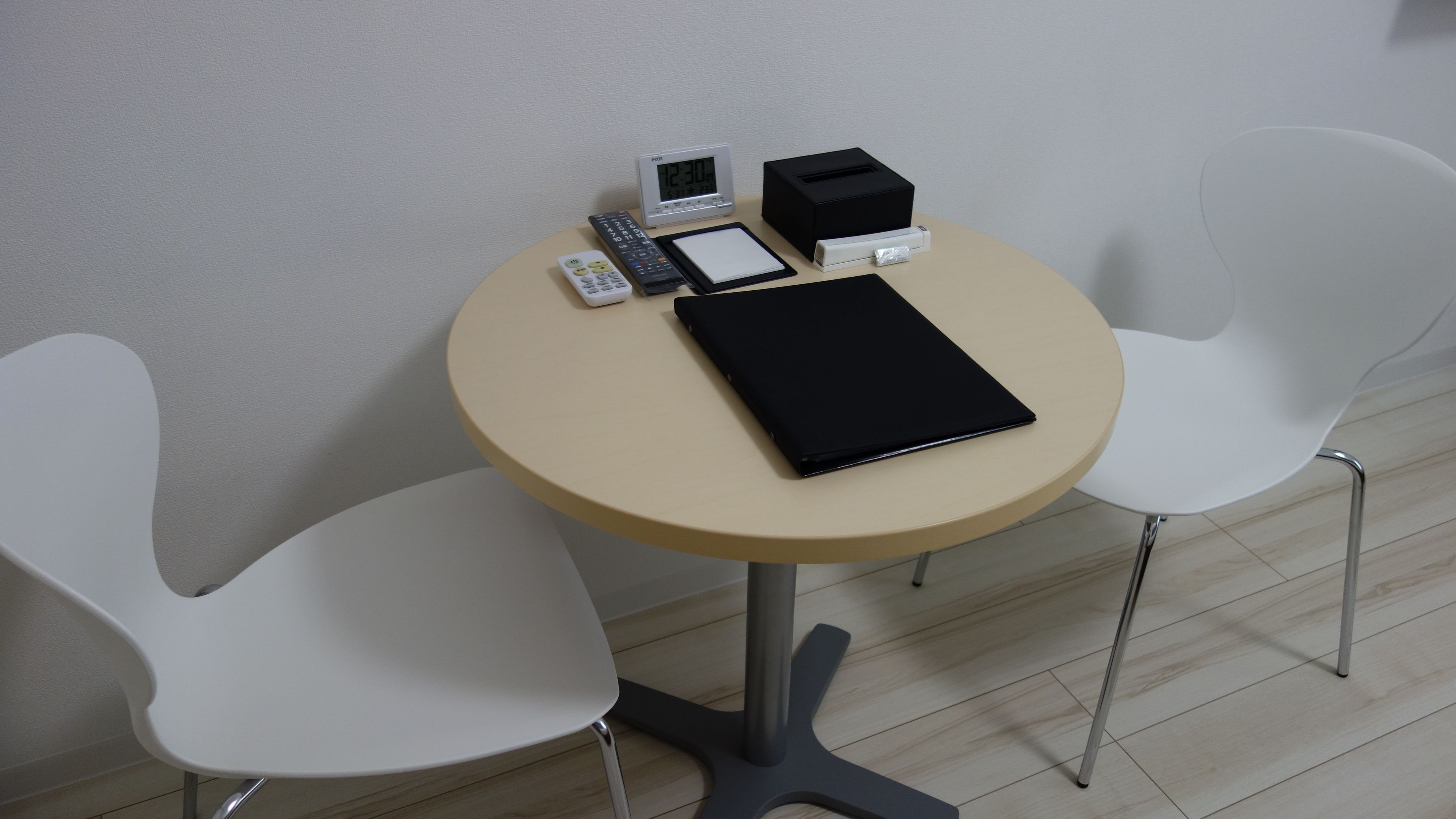 Een kleine ronde tafel met een zwart notitieboek, een witte klok, een afstandsbediening en een zwarte doos. Er staan twee witte stoelen om de tafel heen.