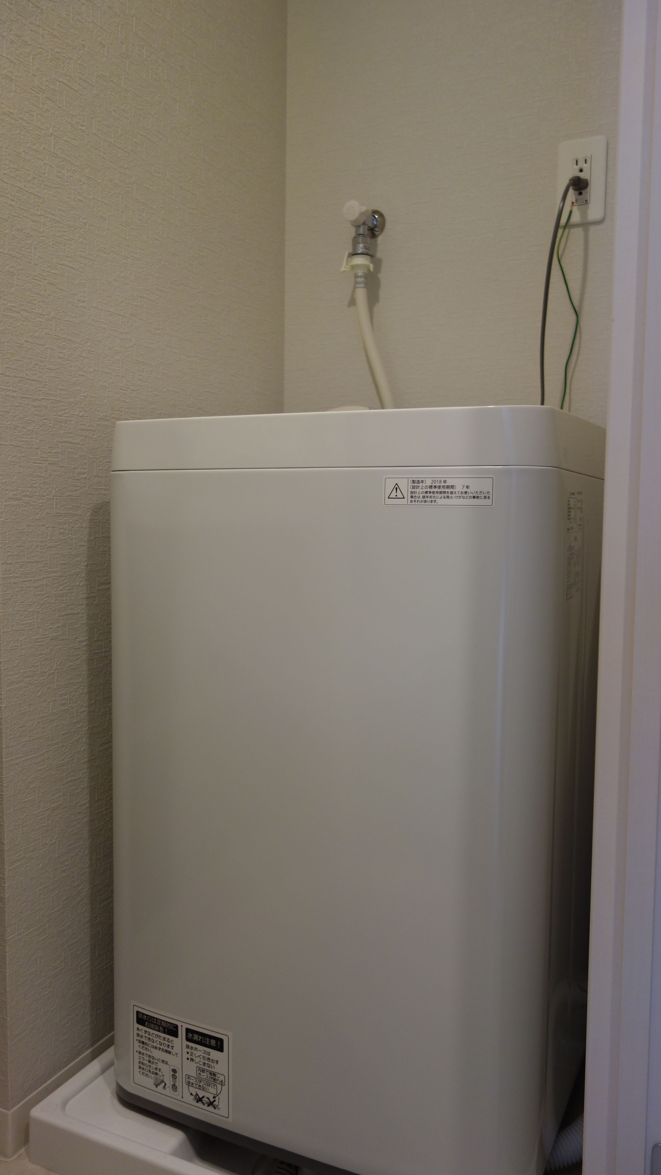 A white water heater in a corner with a power outlet and a cable on the wall.