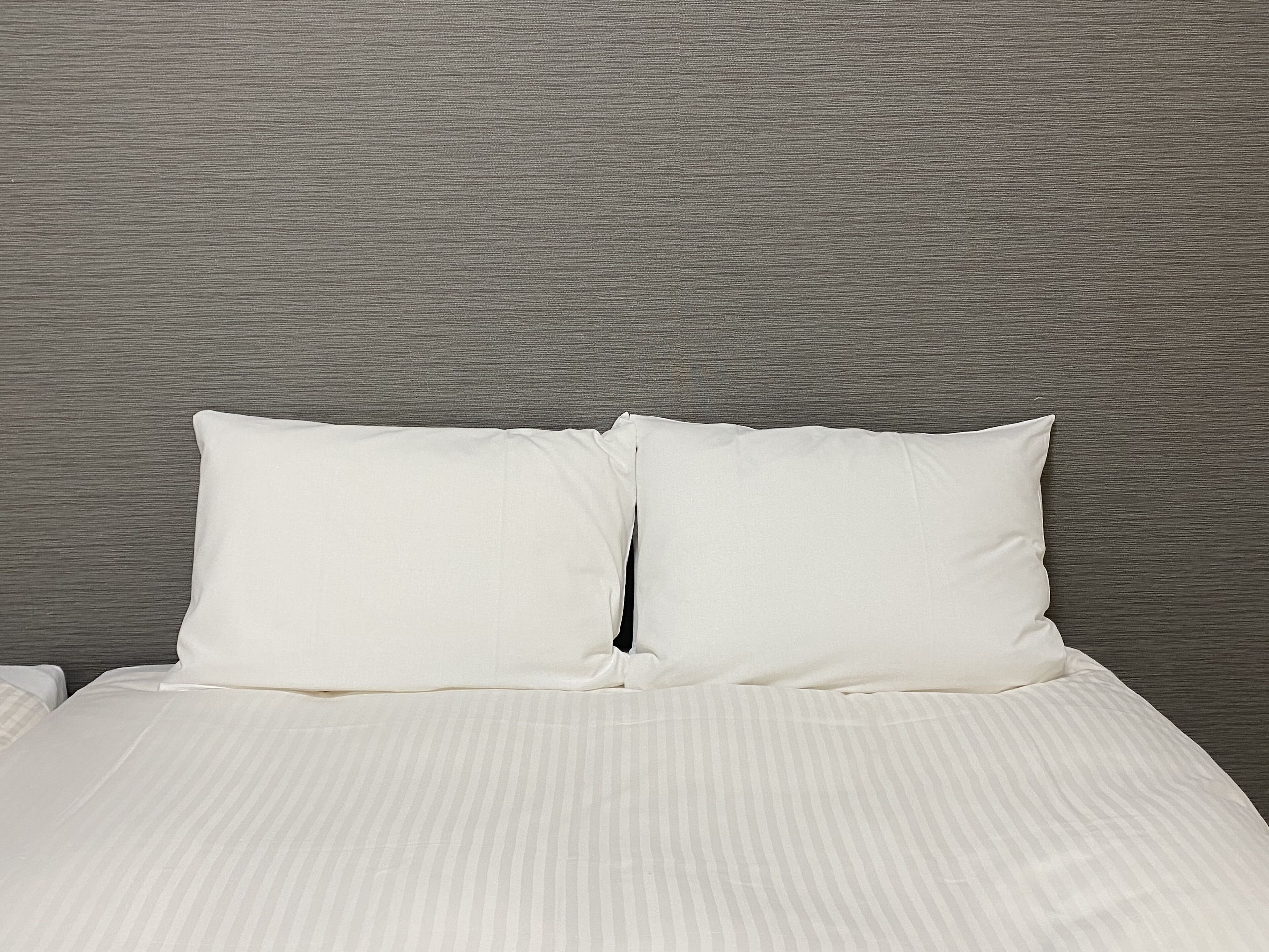 Una cama con ropa de cama blanca y dos almohadas, frente a una pared gris con textura.
