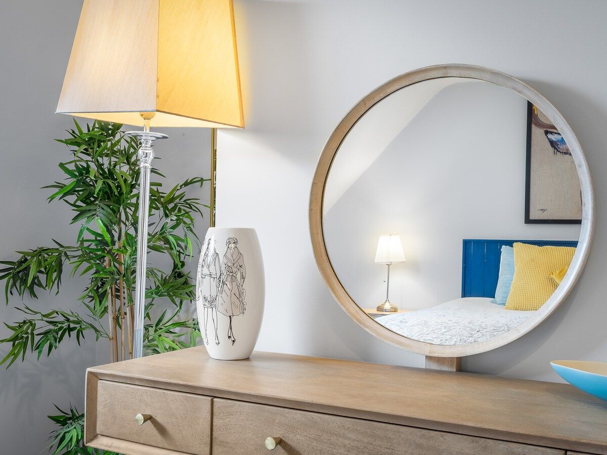 A room with a round mirror, a lamp, a vase with a drawing, and a bed with a blue headboard.