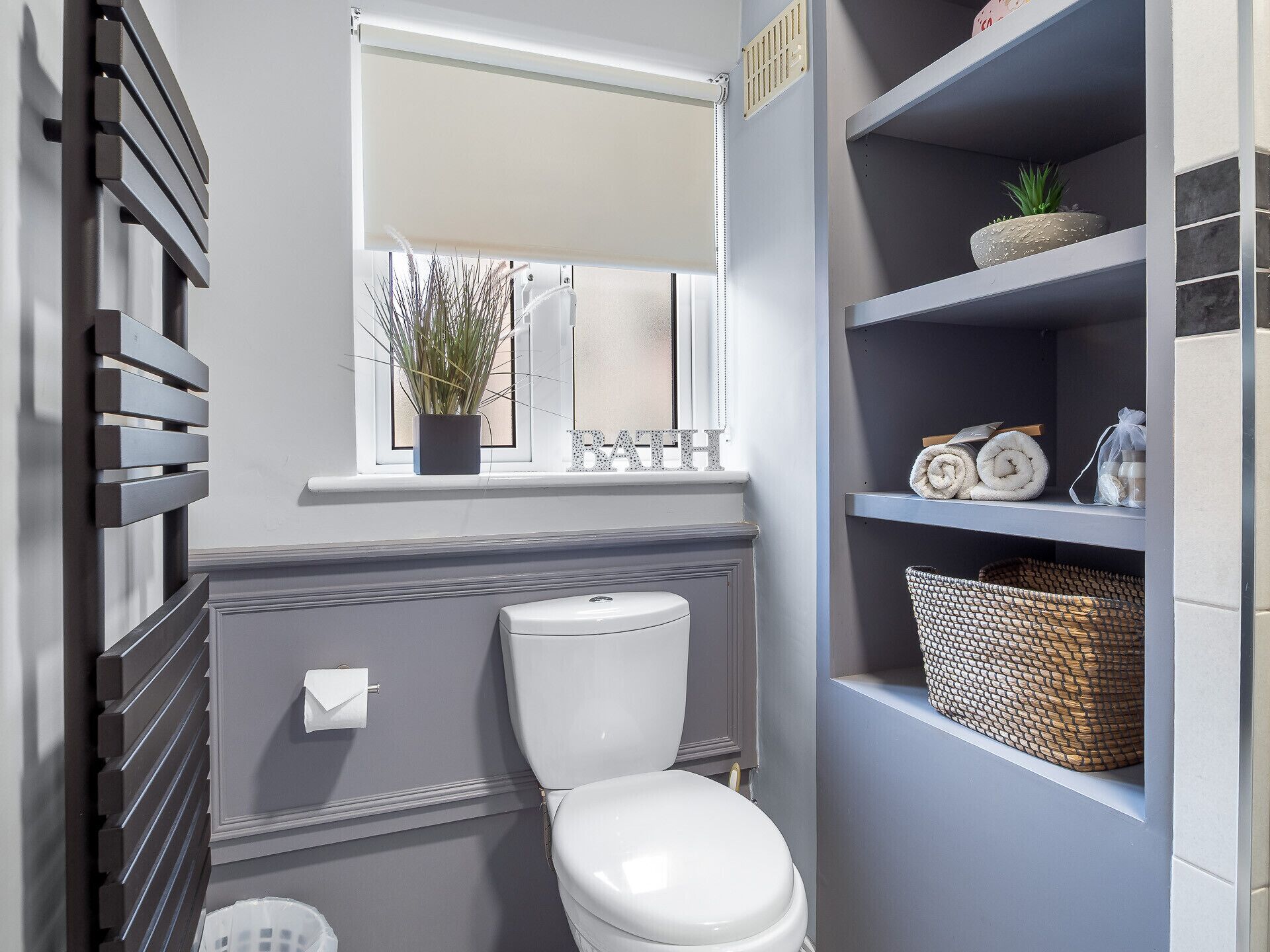 A bathroom with a toilet, a window with a plant, and shelves with towels and baskets.