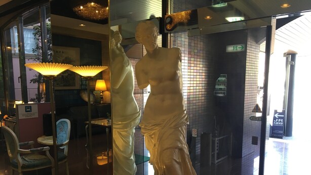 Eine Statue einer Frau in einem Kleid, eine Lampe, ein Stuhl und eine geflieste Wand mit einem Ausgangsschild.