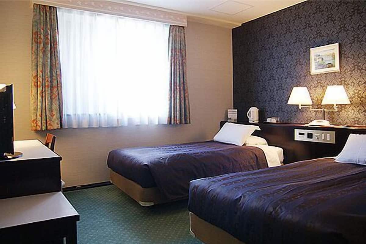 A hotel room with two beds, a desk, a chair, a TV, a window with curtains, and a wall-mounted lamp.