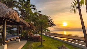 A beachside resort with a thatched-roof structure, a grassy area with stepping stones, and a view of the sunset over the ocean.