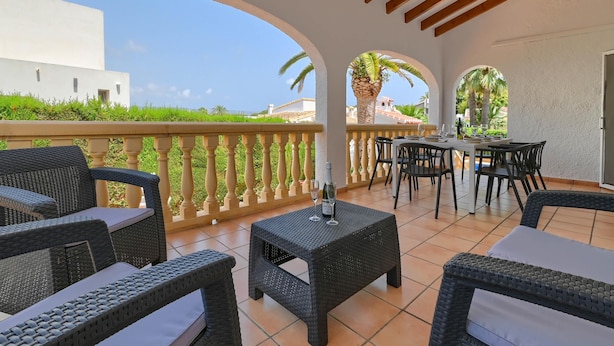 A covered patio area with wicker furniture, a table with two glasses, and a view of a white building and palm trees. - Classic and comfortable villa in Javea, on the Costa Blanca, Spain  with private pool for 6 persons (Javea)