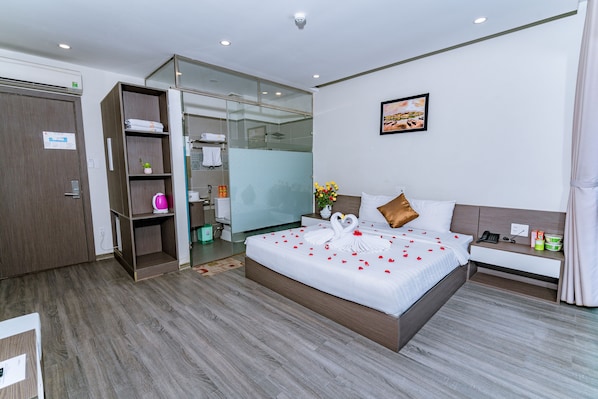 A modern hotel room with a large bed, a walk-in closet, a bathroom with a glass shower, and a small bedside table with a phone and a lamp.