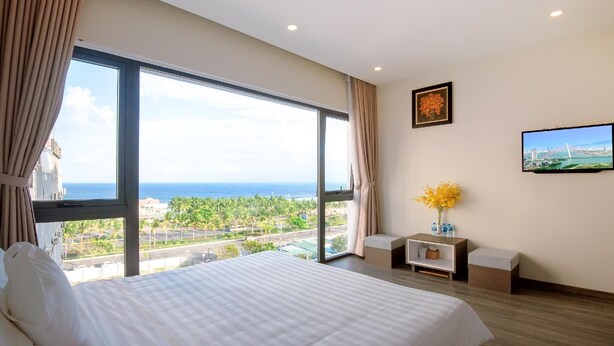 A hotel room with a large bed, a TV, a small table with a vase of flowers, and a view of the ocean and cityscape. - The Shann Hotel Da Nang (Da Nang)