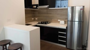 Een compacte keuken met een roestvrijstalen koelkast, een gasfornuis en een klein aanrecht. Op het aanrecht staan twee krukken en een magnetron.