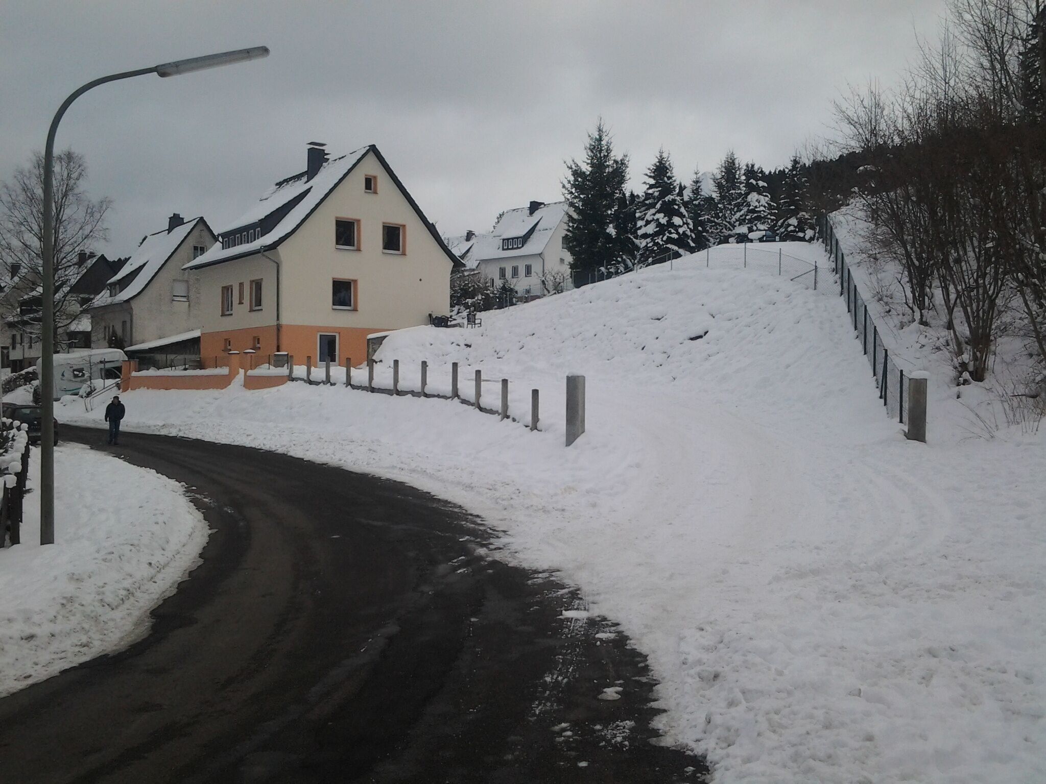 Una calle residencial cubierta de nieve, con casas, una persona caminando y una cerca.