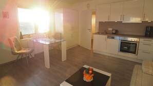 Eine moderne Küche mit weißen Schränken, einem eingebauten Backofen und einer Kaffeemaschine. Es gibt einen Essbereich mit einem Glastisch und Stühlen.