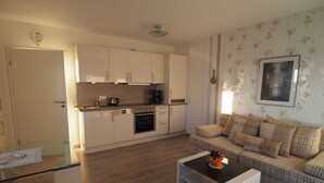 Eine moderne Küche mit weißen Schränken, einer Kaffeemaschine und einem Backofen. Ein Glastisch und eine Couch mit gemusterten Kissen sind ebenfalls vorhanden.