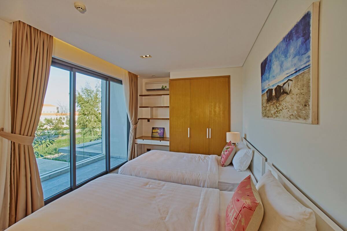 A hotel room with two beds, a large window with curtains, a wooden wardrobe, a painting of a beach, and a small shelf with a picture frame.
