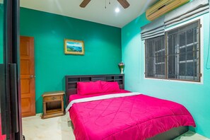 A bedroom with teal walls, a wooden door, a bed with a red bedspread, a nightstand, a small table with a vase, and a window with bars.