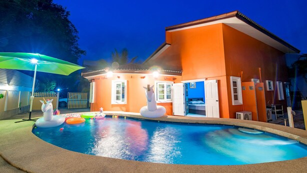 A swimming pool with inflatable unicorns, a covered area with a green umbrella, and a building with orange walls and white doors.