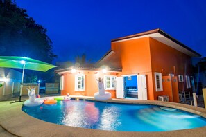 A swimming pool with inflatable unicorns, a covered area with a green umbrella, and a building with orange walls and white doors. - Bann Candy Hua Hin Pool Villa (Hua Hin)