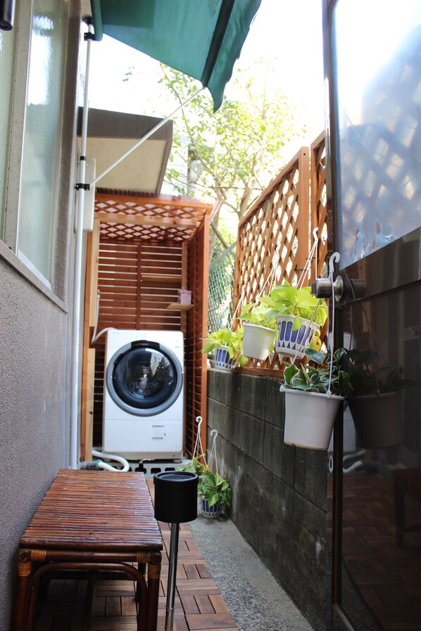 Un espacio exterior reducido con lavadora, mesa de madera y plantas colgantes en macetas.