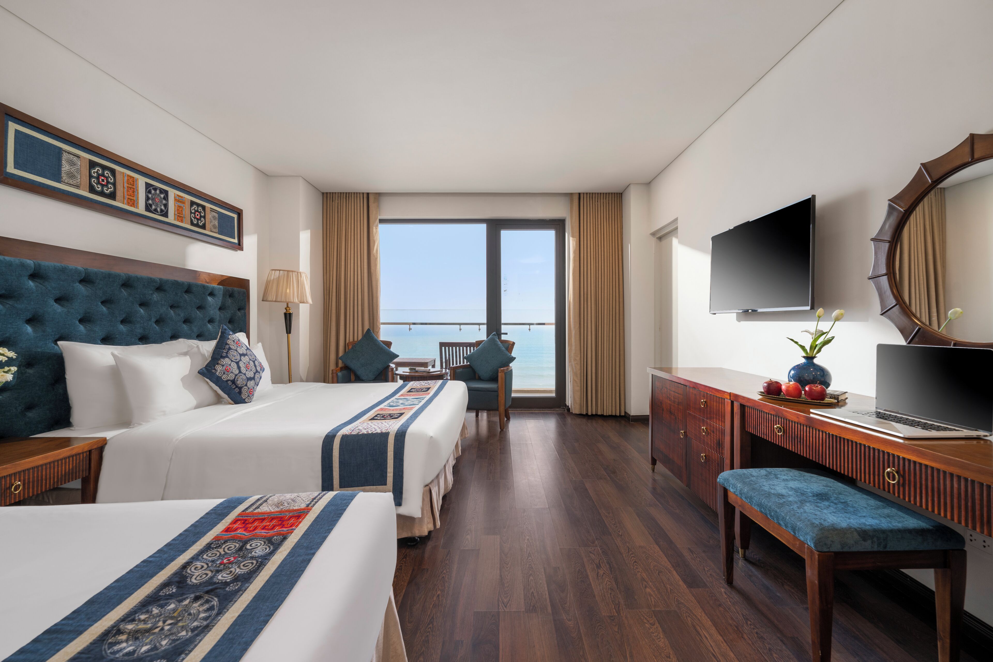 A hotel room with two beds, a desk, a TV, and a view of the sea.