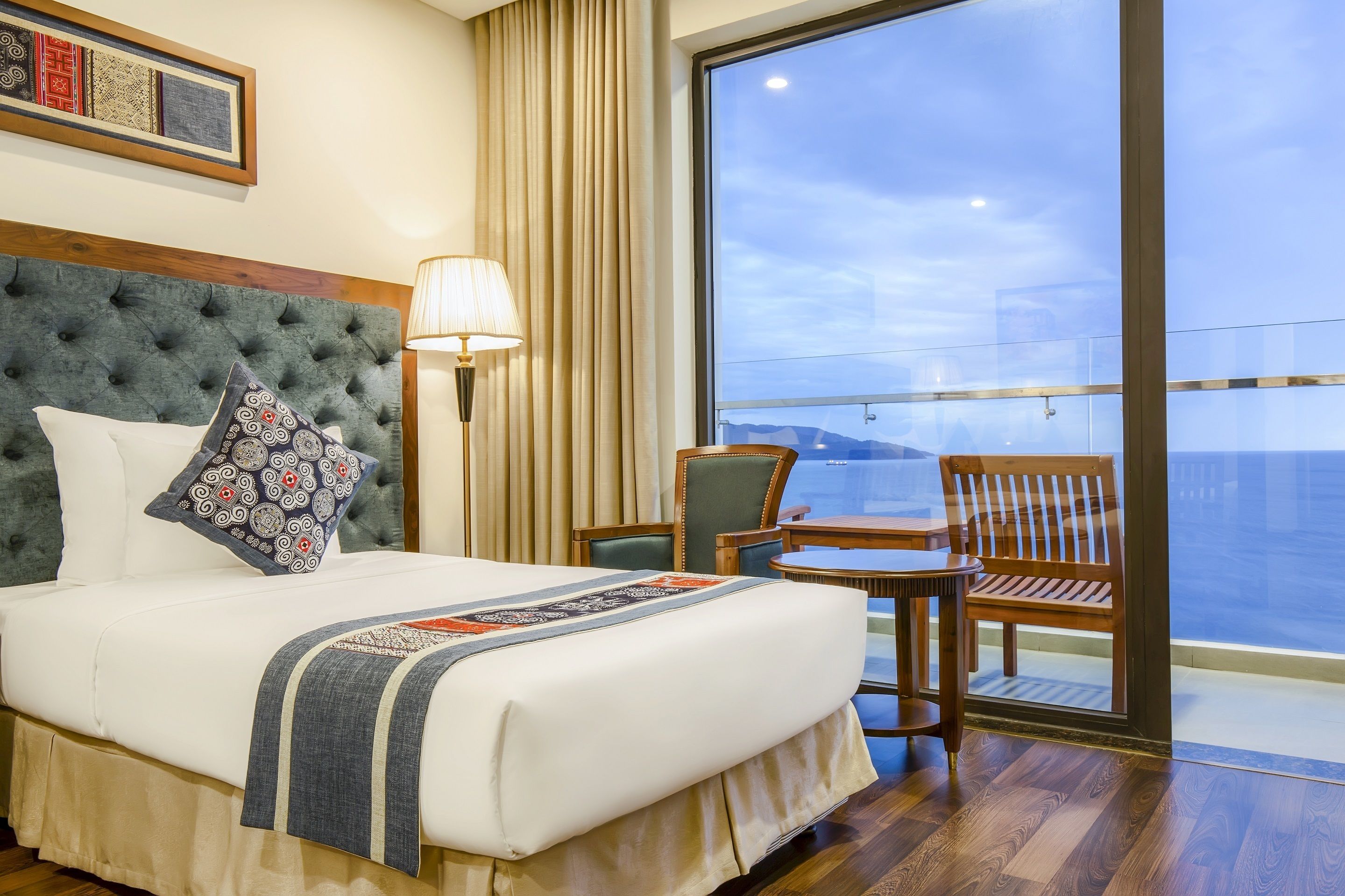 A hotel room with a large bed, a view of the sea, a balcony with a table and chairs, and a large window.