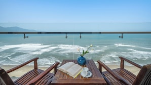 Una mesa de madera con un libro, un jarrón con flores y una taza frente a una playa con vista al océano.