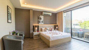 A modern hotel room with a large bed, a bedside table, a chair, a small table with a vase, and a balcony with a view. - Binh Duong Resident Hotel (Da Nang)
