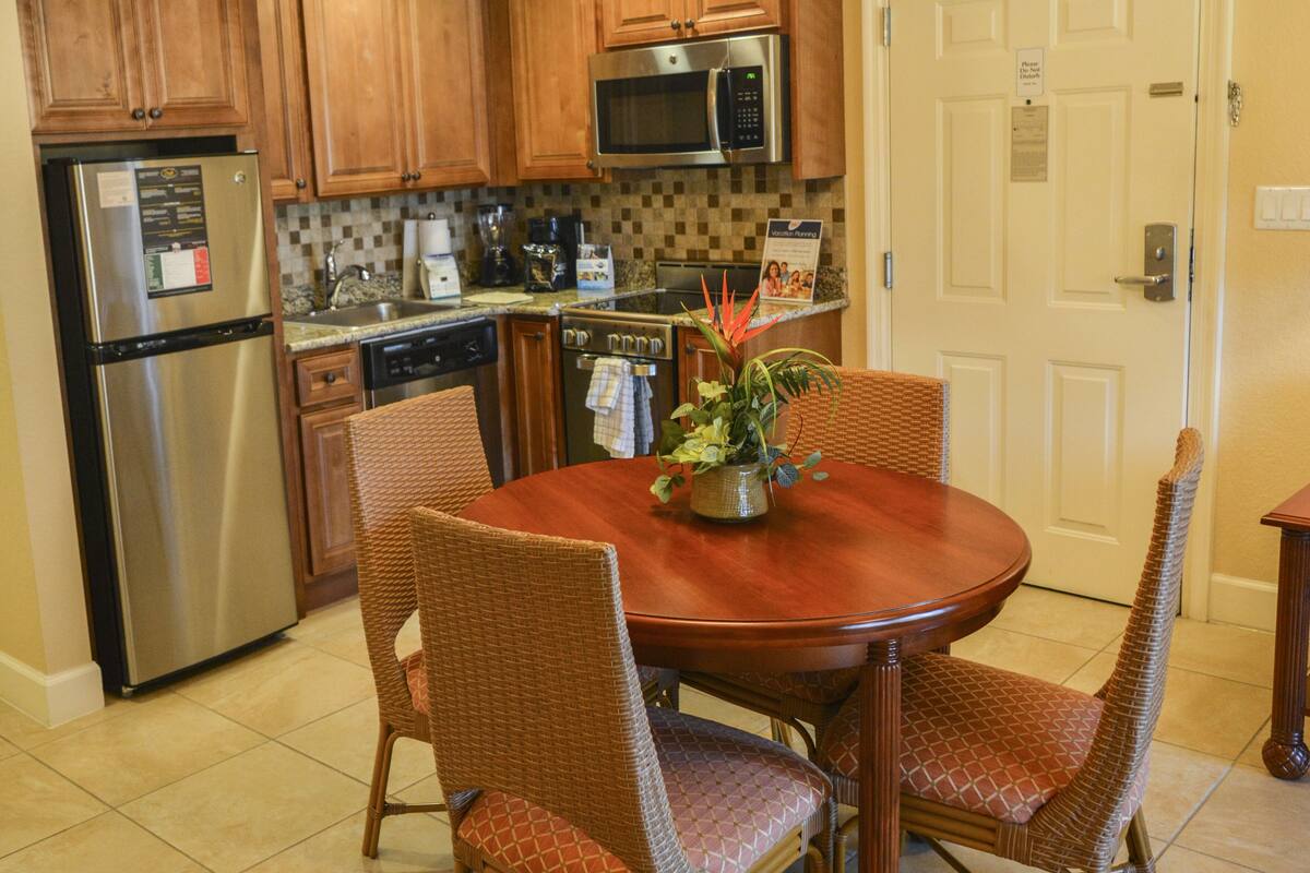 A kitchen with wooden cabinets, a stainless steel refrigerator, a microwave, and a dining table with wicker chairs.