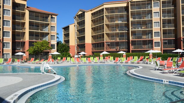 A swimming pool with multiple lounge chairs and umbrellas, surrounded by multi-story buildings with balconies. - Orlando Condo (Orlando)