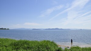 A calm lake with a person standing in the water, surrounded by greenery and distant mountains under a clear sky. - Kanade Fushimiinari (Kyoto)
