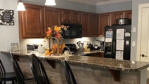 A kitchen with wooden cabinets, a granite countertop, a refrigerator, a microwave, and a bar area with chairs. - Scenic Mountain View, Comforts of Home, Footsteps from Downtown Gatlinburg (Gatlinburg)