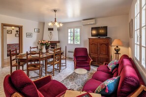 A dining area with a wooden table and chairs, a chandelier, and a television on a cabinet. - Les Lauriers - 8 pers. Single-storey family cottage with enclosed garden - Swimming pool (La Douze)