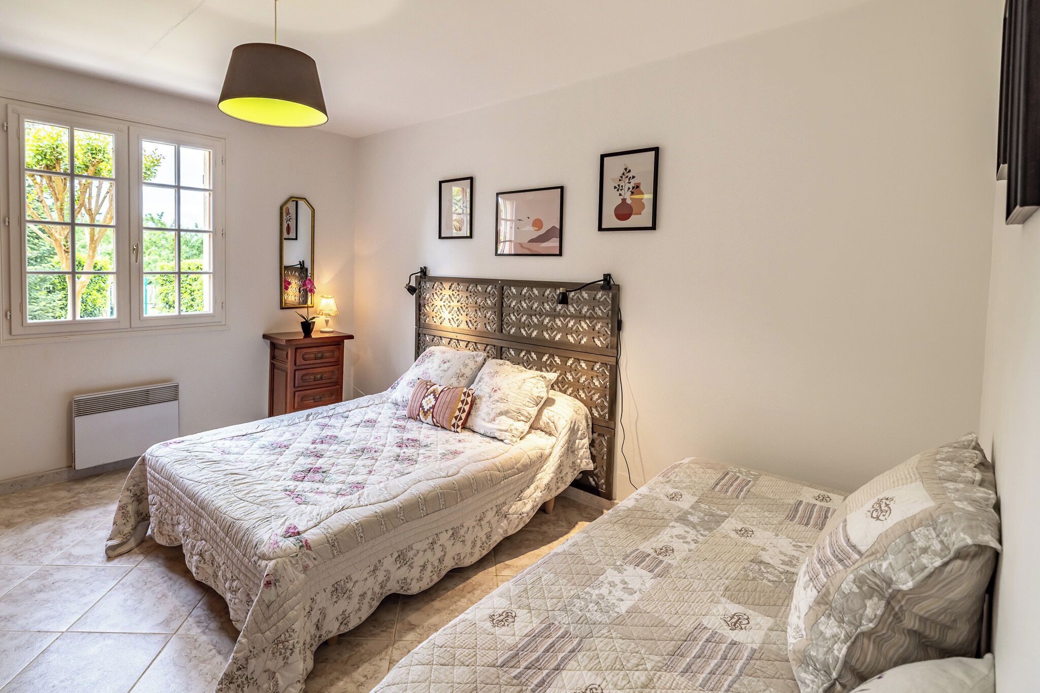 Ein Schlafzimmer mit Bett, Nachttisch, Lampe, Fenster und Wandbildern.