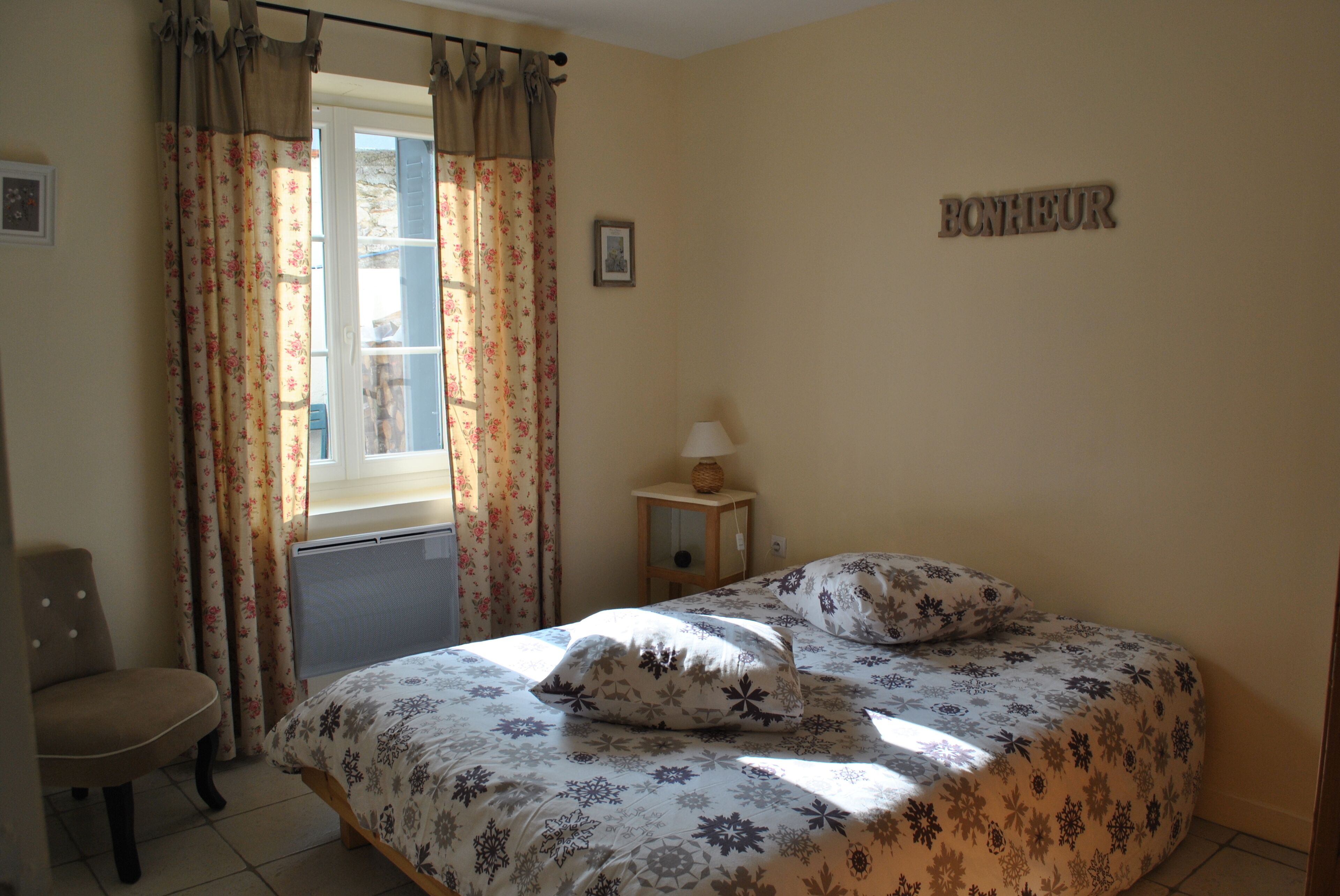 A bedroom with a bed, floral curtains, a chair, a bedside table with a lamp, and a wall sign.