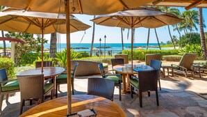 Eine Außenpatio mit runden Tischen und Stühlen, Sonnenschirmen und Blick auf das Meer.