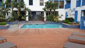 Un espace piscine doté d’une terrasse en bois, entouré de plantes en pot et bordé d’un bâtiment blanc aux ornements bleus.