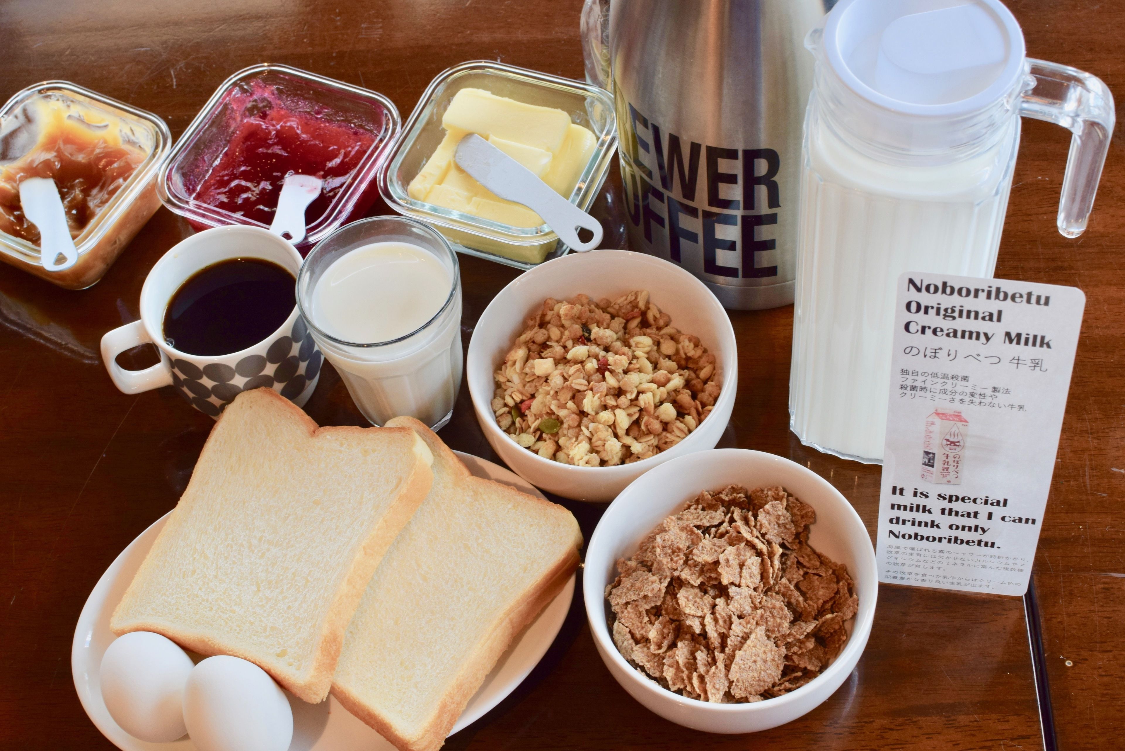 A table with various breakfast items including bread, eggs, cereal, milk, butter, and a coffee maker.
