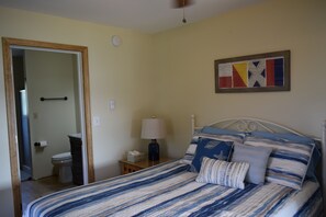 Une chambre à coucher avec un lit, une table de chevet, une lampe, un tableau, et une salle de bain visible à travers une porte ouverte.