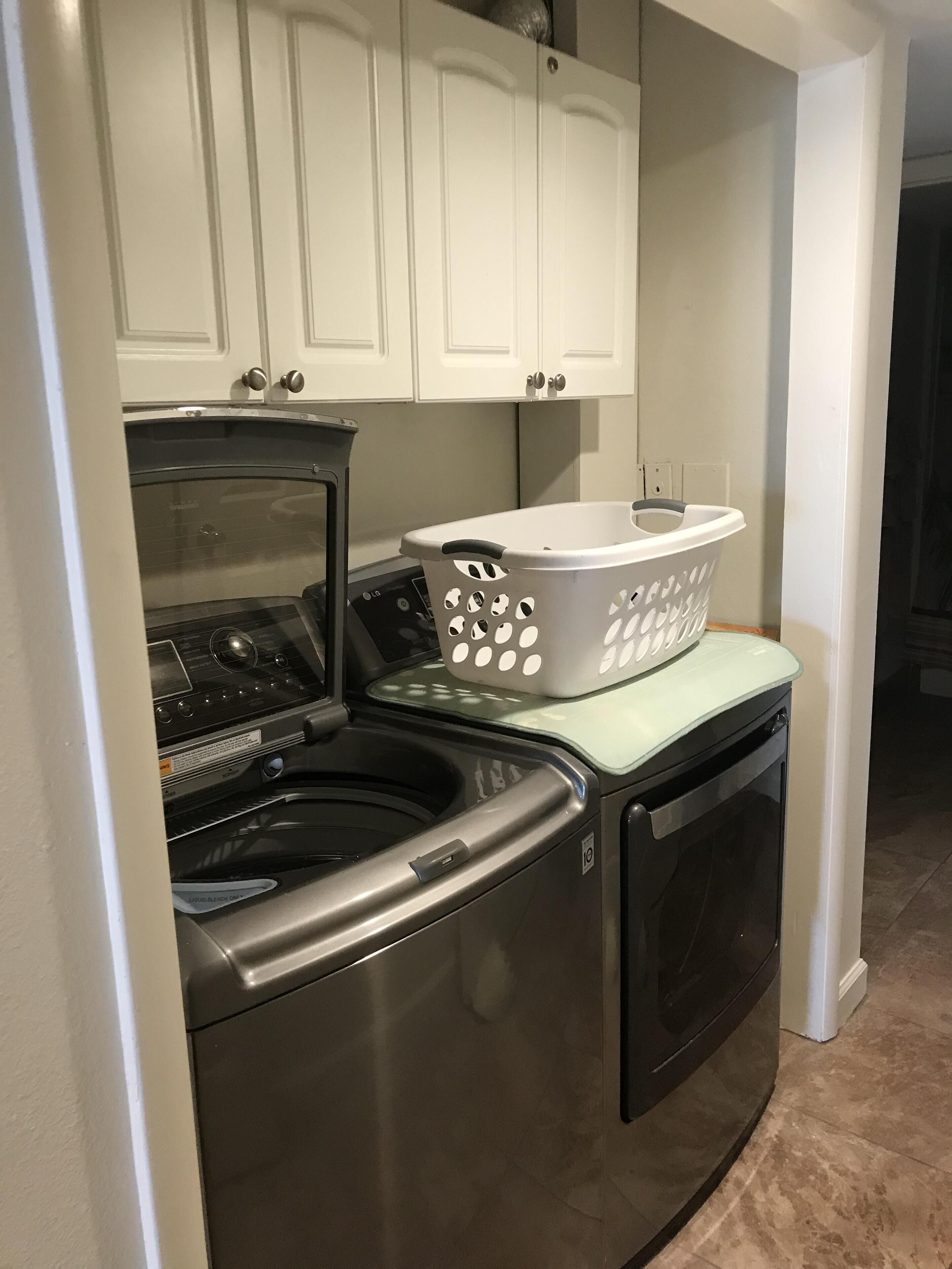 A compact laundry area with a front-loading washer and dryer, a white laundry basket, and white cabinets above.