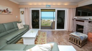 Ein Wohnzimmer mit einem grünen Sofa, einem weißen Couchtisch, einem Babybett und einem Kamin. Es gibt einen großen Fernseher und einen Balkon mit Meerblick.