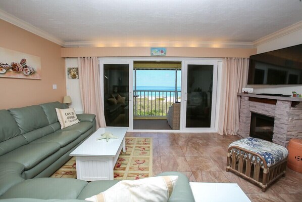 A living room with a green sofa, a white coffee table, a baby crib, and a fireplace. There is a large TV and a balcony with a view of the sea. - Direct Beachfront Gulf with Amazing Views (Longboat Key)