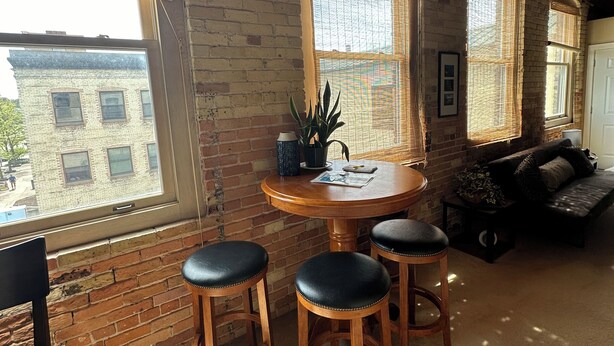A cozy room with a brick wall, a round wooden table with stools, a sofa, and large windows with blinds. - Fantastic Penthouse Downtown Grand Haven! Harbor Front Views! Walk Everywhere! (Grand Haven)