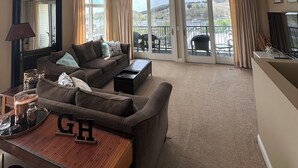 A spacious living room with a sofa, coffee table, and a dining area with a wooden table and letters "GH". Large windows offer a view of the outdoors. - Fantastic Penthouse Downtown Grand Haven! Harbor Front Views! Walk Everywhere! (Grand Haven)