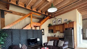 A spacious living room with a large brown leather sofa, a wooden staircase, and a kitchen area with wooden cabinets and a refrigerator. - Fantastic Penthouse Downtown Grand Haven! Harbor Front Views! Walk Everywhere! (Grand Haven)