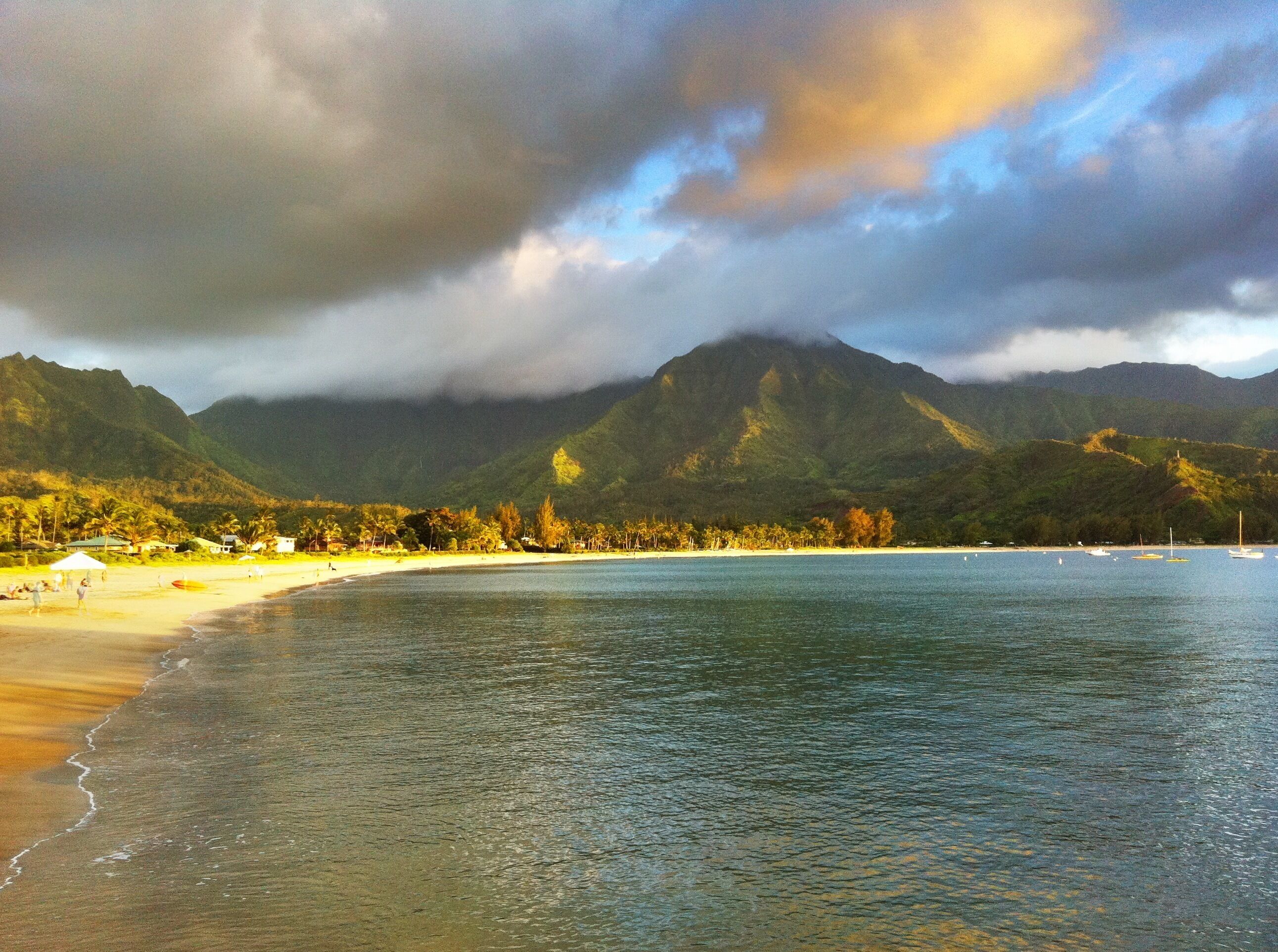 A beach with clear water, a mountain in the background, and a cloudy sky.