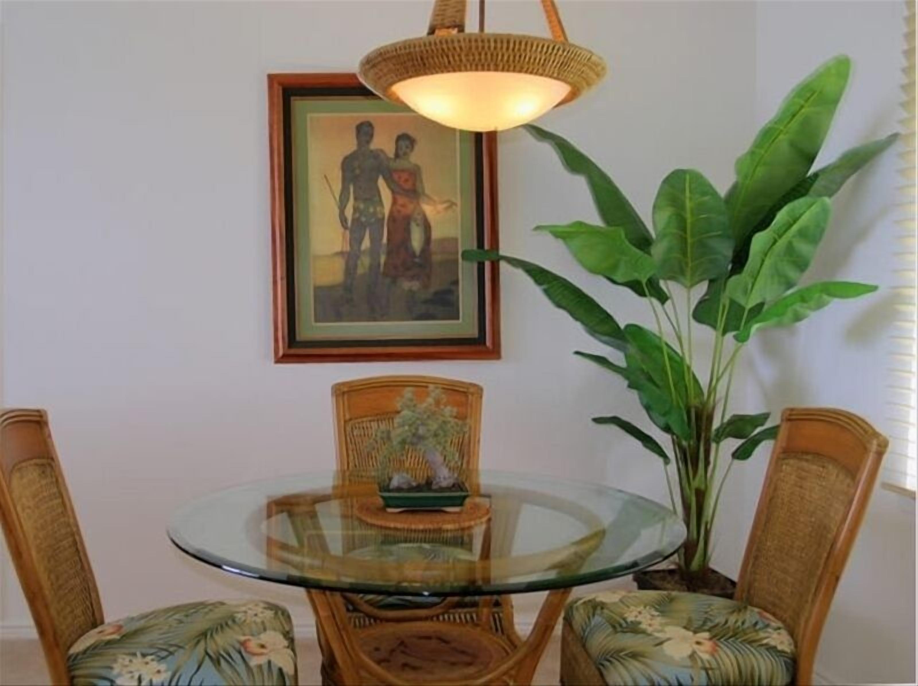 Un área de comedor con una mesa de vidrio rodeada de sillas de mimbre, un cuadro en la pared y una planta verde grande.