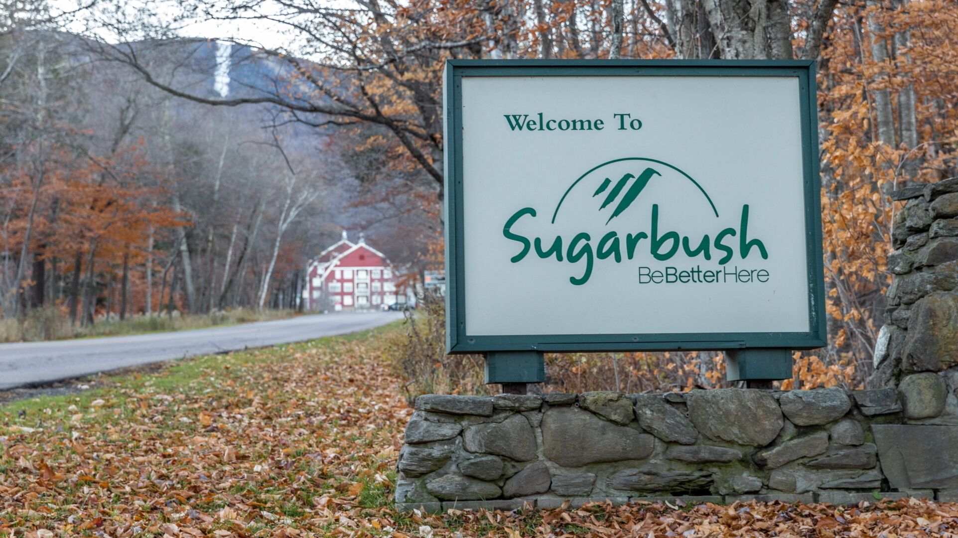 A welcome sign for Sugarbush with a stone wall and autumn leaves.