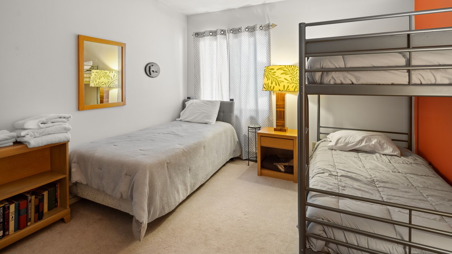 A compact hotel room with a bunk bed, a single bed, a bedside table with a lamp, a mirror, a bookshelf, and a window with curtains.