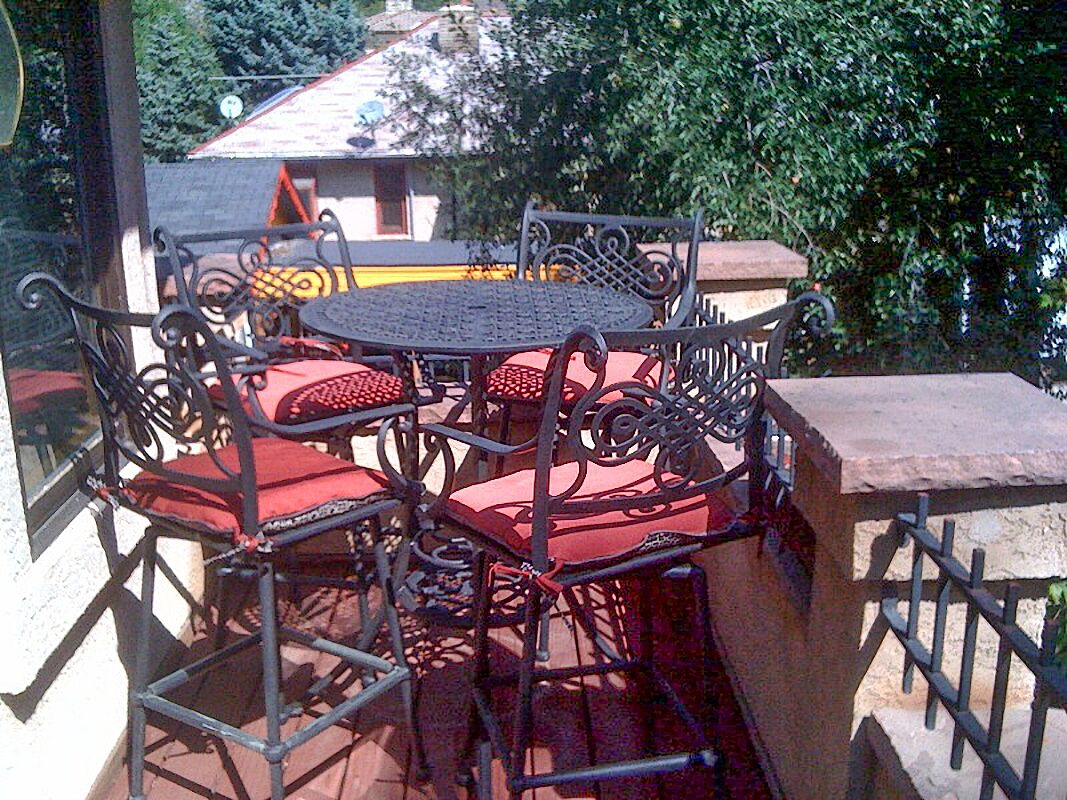 Een ronde metalen tafel met rode fauteuils, geplaatst op een balkon met uitzicht op bomen en een gebouw op de achtergrond.