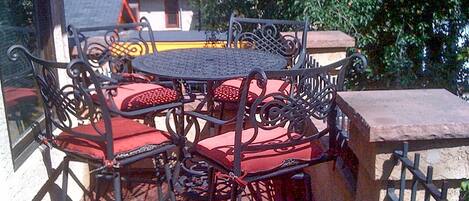 Een ronde metalen tafel met rode fauteuils, geplaatst op een balkon met uitzicht op bomen en een gebouw op de achtergrond.