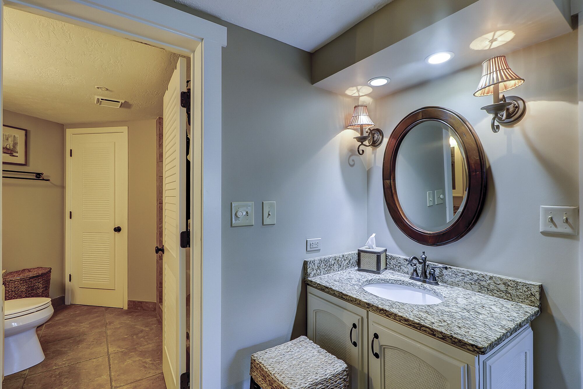 A bathroom with a white toilet, a sink with a marble countertop, a wicker stool, and a mirror with a wooden frame.