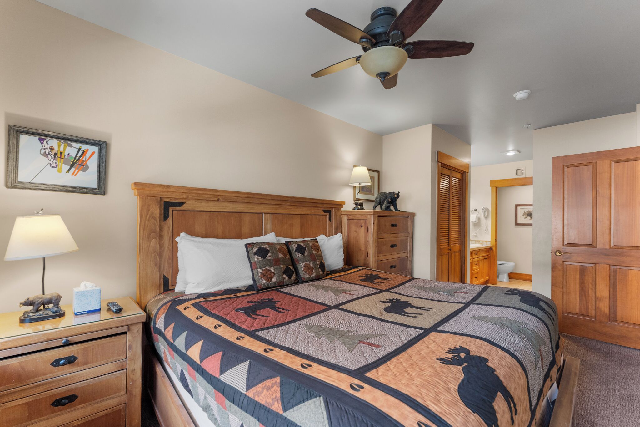A bedroom with a wooden headboard, a quilt with animal prints, a nightstand with a lamp, and a ceiling fan.